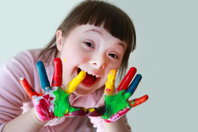 A young light-skinned girl with down syndrome smiling into the camera. Multicolored paint covers her hands
