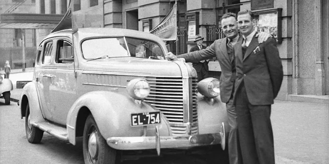 Black and white photograph of Sir Andrew Bruce Small standing next to Sir Hubert Oppy Opperman. There is a car sitting next to the men. 