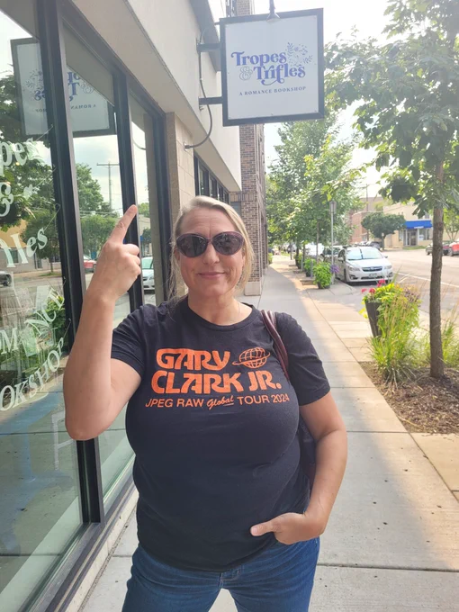 Amys Farstrider standing under the Tropes & Trifels book store sign, pointing to the sign with her right hand. Trees, cars, and the road in the background.  