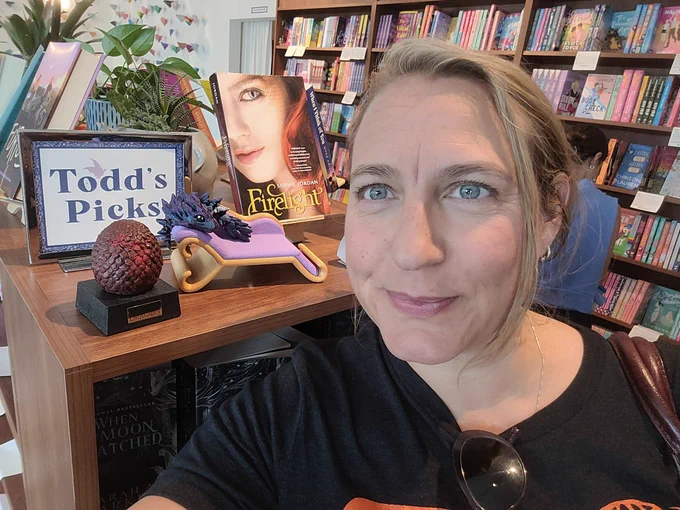 Amys Farstrider taking a selfie in a book store with a large bookshelf in the far back and standing in front of the shelf labeled "Todd's Picks" showcasing Todd the dragon on his chaise lounge.