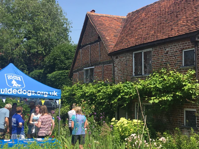 This image from a previous Guide Dogs open day at Milton's Cottage shows the red brick, timber-frame front of Milton's Cottage surrounded by a green vine and the greenery of the surrounding garden. Volunteers in blue Guide Dogs t-shirts are chatting to visitors in the shade of a blue Guide Dogs gazebo.