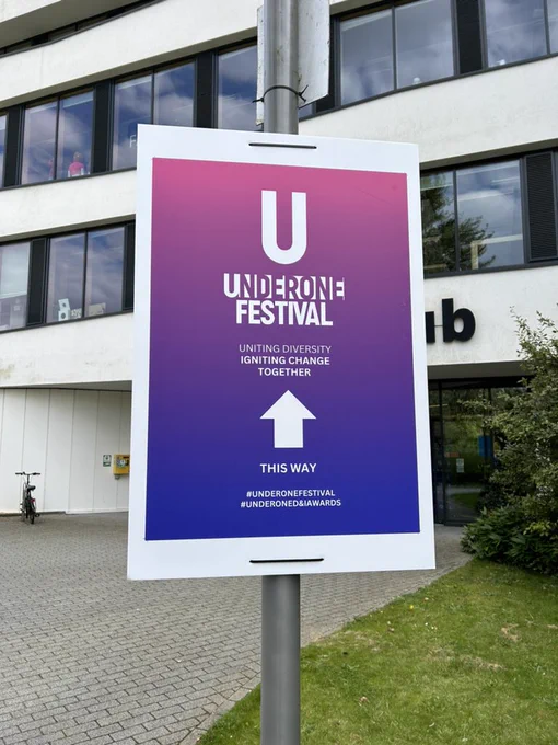 Photograph shows a poster attached to a pole outside the Creative Hub building at the University of Northampton. The poster reads UnderOne Festival. Uniting Diversity. Igniting Change Together. There is an arrow pointing up. Under the arrow reads This Way #UnderOneFestival and #UnderOneD&IAwards