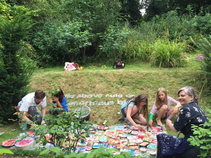 This image shows a previous workshop in the garden at Milton's Cottage with 3 children and 2 workshop leaders surrounded by green plants and lots of colourful painting palettes.