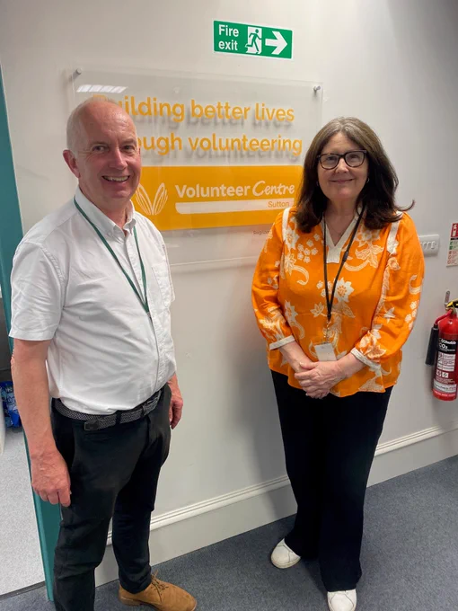 The photo shows Councillor Barry Lewis standing next to Anita Maullin, the CEO of Volunteer Centre Sutton. Cllr Lewis is standing on the right and Anita is standing on the left. Behind them is a sign which says Volunteer Centre Sutton.