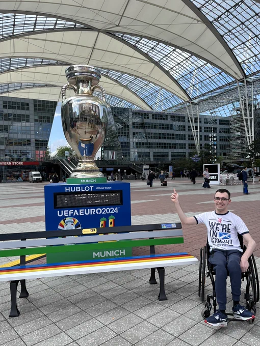 Laurie pictured outside Munich Airport pointing towards a replica of the European Championship trophy next to a Euro 2024-themed bench.