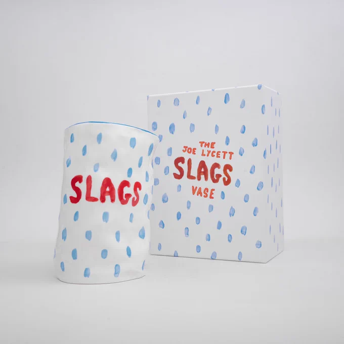 A sloped white ceramic vase with blue dots and the word ‘SLAGS’ written on it, next to a box in the same colours that reads ‘The Joe Lycett Slags Vase’