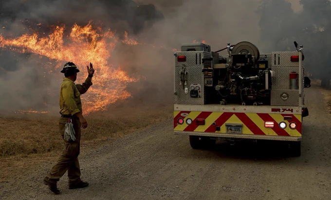 A fire blazes in the background of a firefighter and a firetruck.