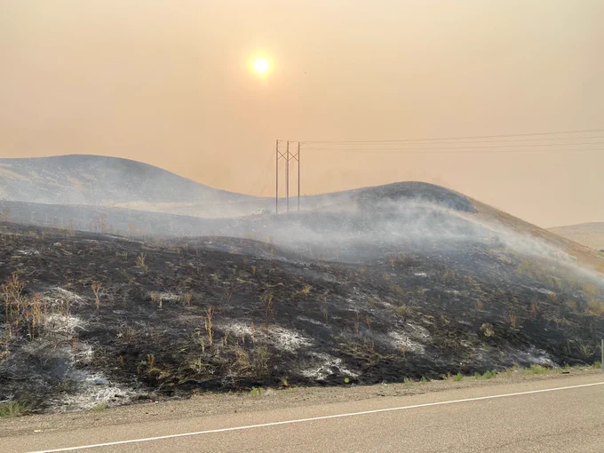 Smoky landscape showing burnt vegetation along Interstate 84 near milepost 346. Hills in the background are under a hazy sky obscured by wildfire smoke.