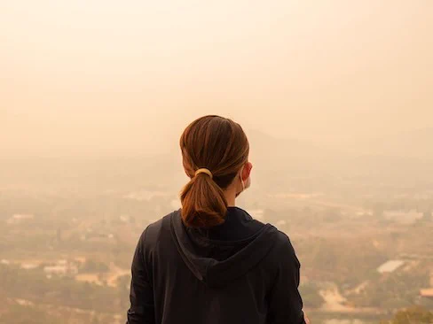 The image shows the back of a person standing and looking out over a hazy landscape, obscuring the view of the landscape below. The overall atmosphere appears to be smoggy or foggy, suggesting poor air quality.