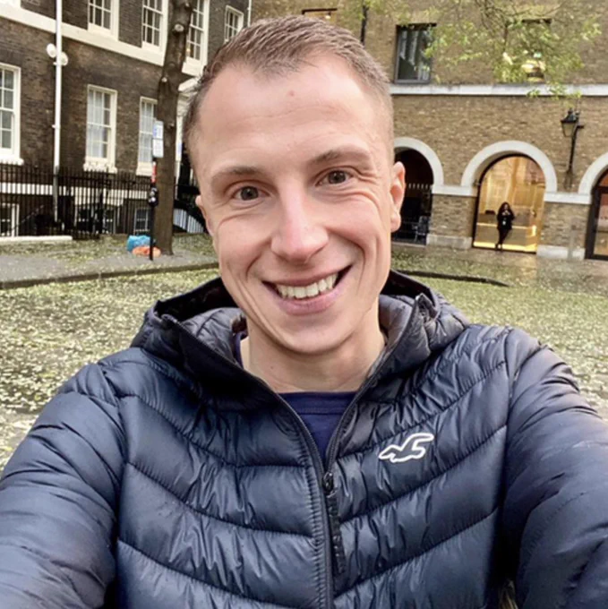 James has short hair and pale skin. He's smiling at the camera, wearing a navy blue puffer jacket. 