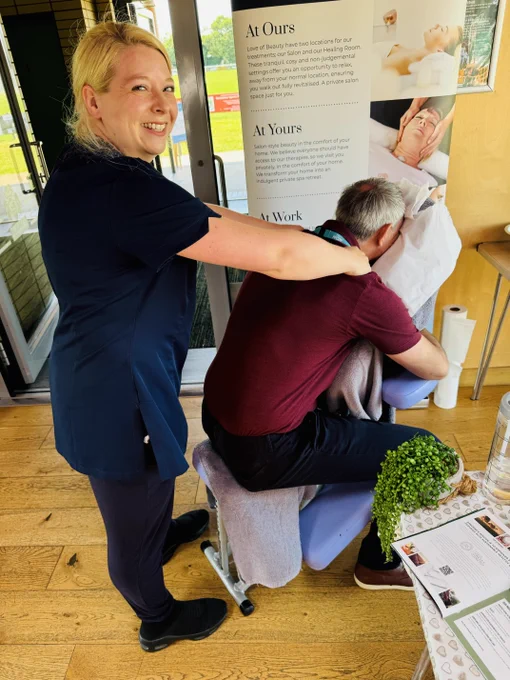 James sitting on a chair leaning forward with his face in a hole. The lady doing the massage is smiling looking at the camera, she has her hands on James neck giving him a shoulder massage. Conflict.