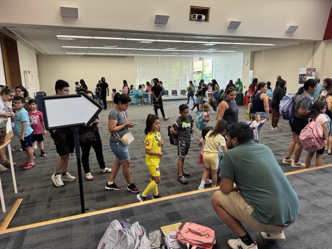 A large group of diverse children standing in line waiting to get a backpack full of school supplies. 