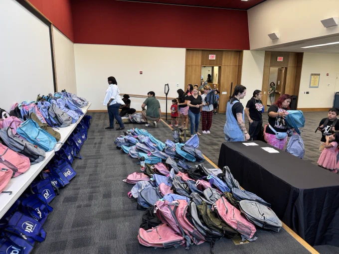 Photo of a stage in the library meeting room. The stage is full of pink, black, blue and purple children's backpacks. 