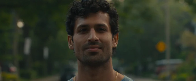 A film still from Shook. A man stares directly into the camera, his face and neck visible. He has dark curly hair and light facial hair and is wearing a blue shirt and a gold chain. He has a slight smile on his face. The background is out of focus but he appears to be standing on a suburban street in spring or summer, as many green trees are visible behind him, as well as some cars and traffic signs.
