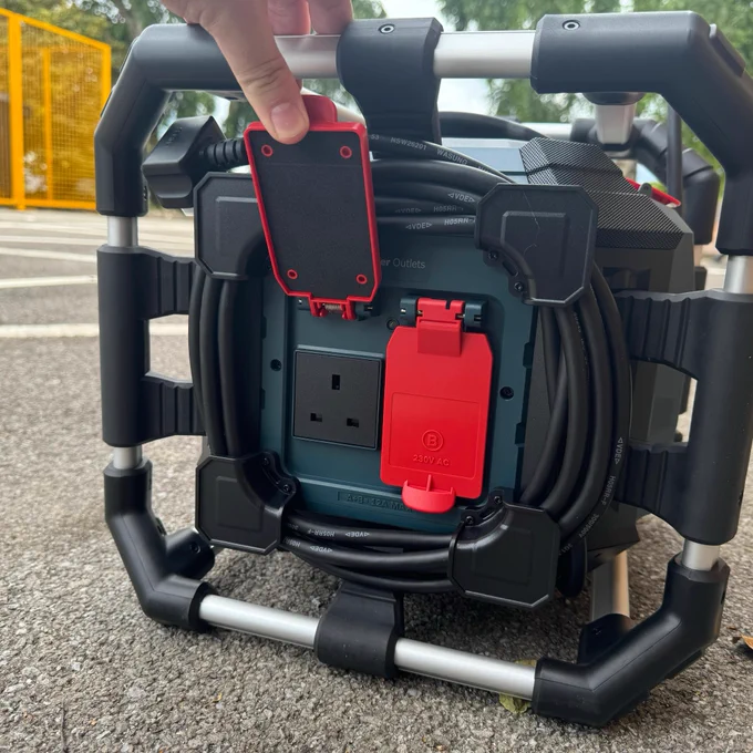 Close-up view of a portable power station with a hand pressing a red reset button. The device features multiple output ports including standard electrical outlets and USB ports, encased in a black frame with a handle.