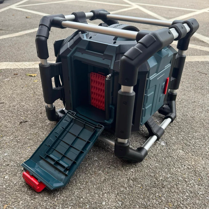 A portable, black and gray radio is placed on an asphalt surface. The back tray is pulled out, showing where the battery is inserted.