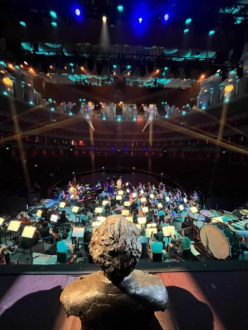 The back of the bust of Henry Wood with orchestra members in front on the stage. The empty seats of the Royal Albert Hall at the back of the photo.