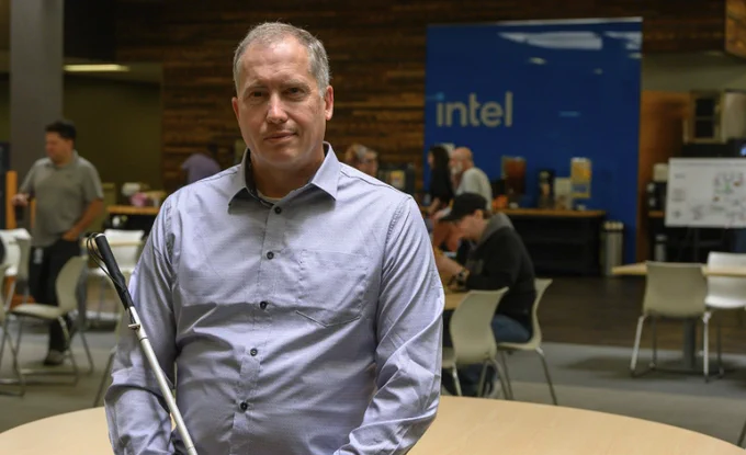 A man with a white cane is standing in a room with a table and chairs. There's an Intel sign in the background.

