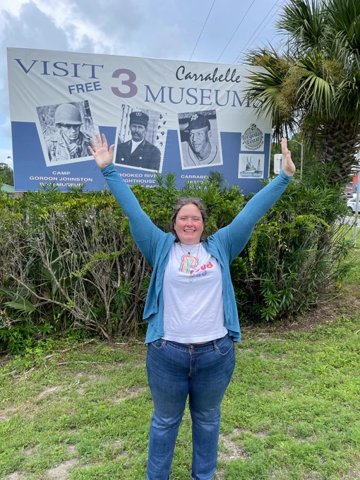 A tall, middle aged white lady with dark blonde hair in long braids has a huge grin on her face as she frames a photo of playing age Buck O’Neil with her outstretched hands by the side of a road. The billboard reads Visit Three Carabelle Museums 