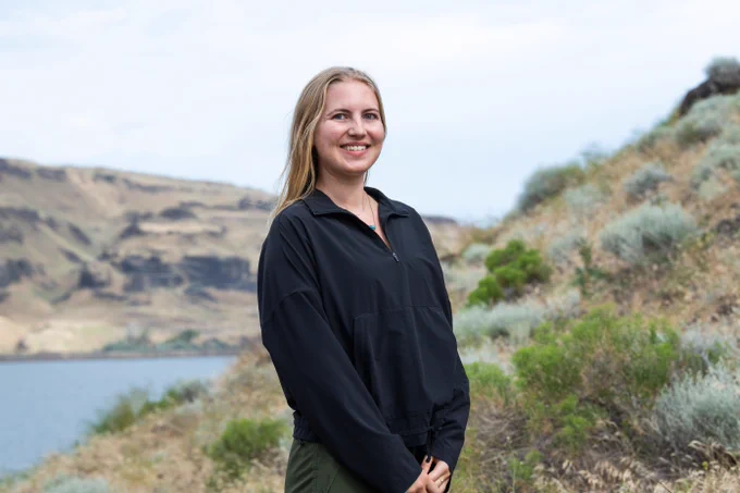 Photo of PNNL intern Madeline Bartels smiling at the camera, with sparsely vegetated hills and the Columbia River in the background. Madeline visited the basalt field near the site of the Wallula, WA carbon storage demonstration project, where in 2015 PNNL researchers validated the rapid CO2 mineralization rates observed from years of laboratory testing with basalts.
