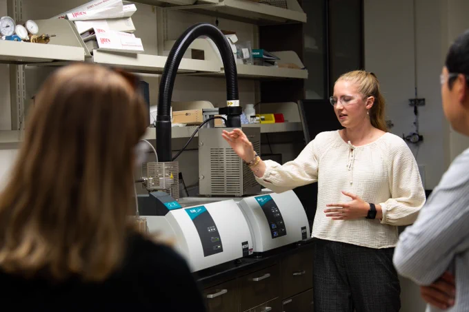 Photo of intern Madeline Bartels standing in a PNNL carbon mineralization lab, demonstrating to visiting researchers the equipment and techniques used to prepare and process samples of CO2 mineralized in basalt.