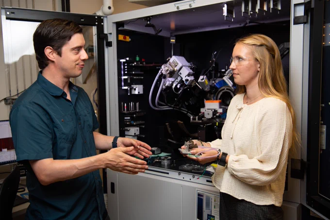 Photo of intern Madeline Bartels holding a rock, standing in front of laboratory equipment, and speaking with chemist Quin Miller from PNNL's Energy and Critical Materials Group.