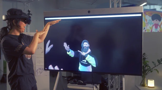 A person wearing VR-glasses gestures toward a large monitor displaying a 3D animated figure, in an office environment with educational posters on the wall.
