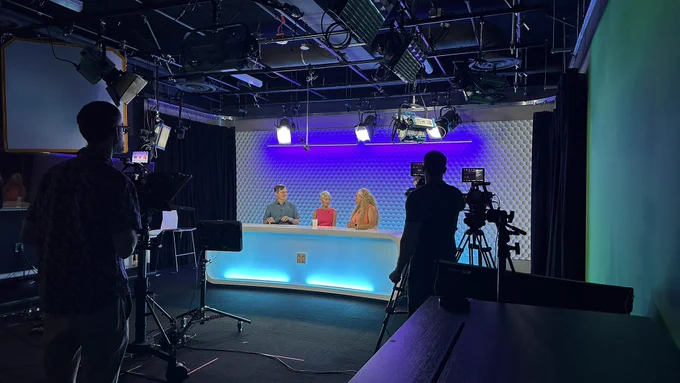 A picture of 3 people sitting at a table in a video recording studio with a blue and purple background