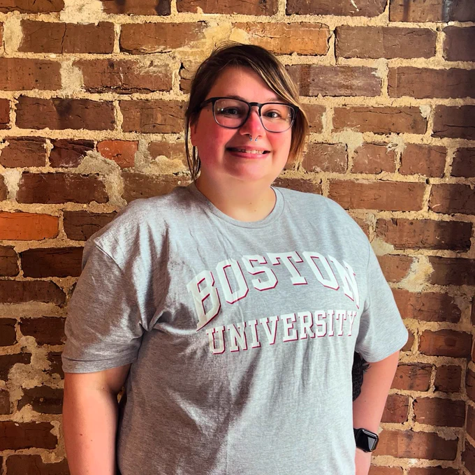 A photo of Micah standing in front of a brick wall. They are wearing a grey t-shirt with “Boston University” written on the front.
