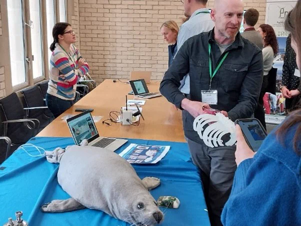 The image shows an event where numerous people are gathered around exhibition tables. On the tables are laptops, equipment and a toy seal. 