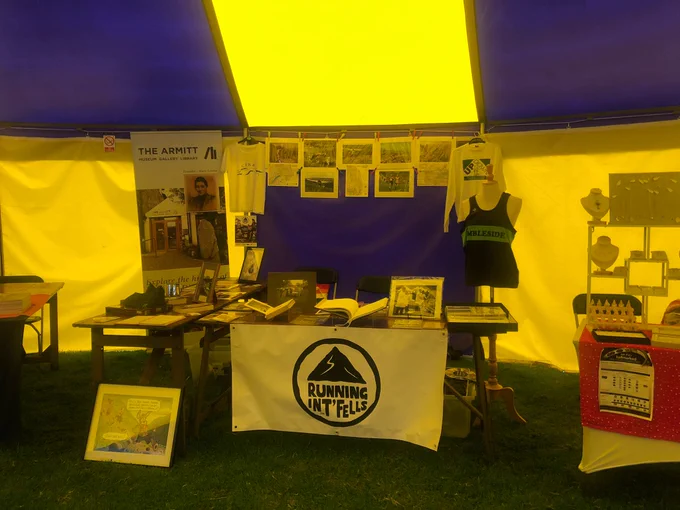 The Running Int Fells stall in the main tent at Ambleside Sports. The stripey tent is yellow and purple, bathing the stall in those colours. The table is covered with books and objects, with photos on display on the canvas tent wall behind it. A large banner for the museum is on the left hand side of the table. A large white banner with the black Running Int Fells logo is on the front