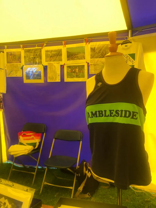 A black running vest with Ambleside written on the front is displayed on a mannequin at the Running Int Fells stall. There are photos of fell runners pegged onto a string on the tent wall behind the mannequin