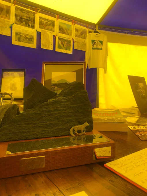 A close up of a slate and metal fell running trophy mounted on wood on the stall table. A small metal wolf stands in front of slate mountains, with a metal plaque at its feet as a lake