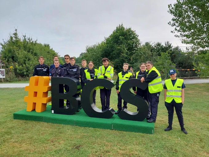 Cadets and cadet leaders stood in a field behind a large sign displaying #BCS
