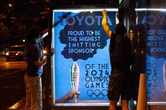 Two people install an artwork in a bus shelter. The artwork shows an olympic torch billowing smoke and the words "Toyota. Proud to by the highest emitting sponsor of the 2024 Olympic Games"