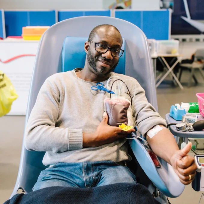 A man sits in a donor chair holding a bag of his donated blood. He is smiling and is doing a thumbs up. 
