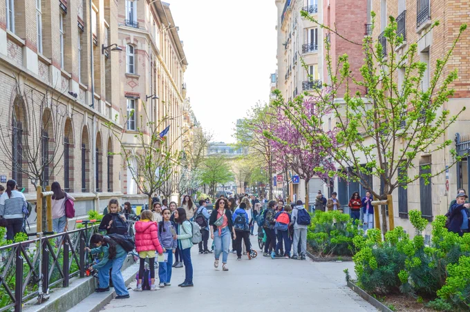 A car-free “school street” in Paris filled with playing children includes bike parking, vegetated areas, blossoming trees, and white asphalt painted with colourful games.