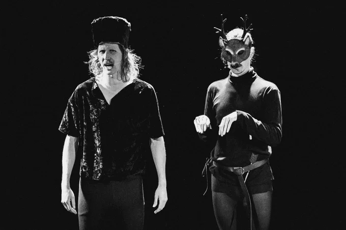 Black and white photo of two men; one is wearing a large wool hat, the other wears a deer-shaped mask.