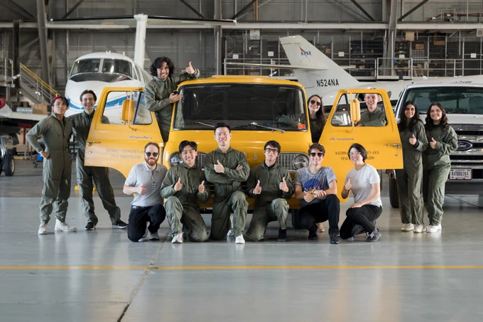 Interns wearing green flight suits stand in front of and beside a yellow van in the flight research hanger at NASA Langley. Many are giving thumbs up. 