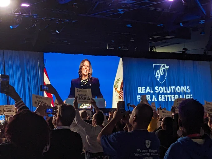 In a large convention hall, Vice President Kamala Harris appears on a projector screen standing at a podium and smiling. On the curtain besides the screen is projected the AFT logo and "Real Solutions for a Better Life". A large crowd watches holding signs that read "AFTVotes Kamala Harris for President".