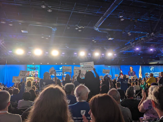 A large crowd stands assembled in a large convention hall. Vice President Kamala Harris stands at the front of the room under the words "Vice President Kamala Harris". She also appears larger on a projector screen standing and smiling. On the curtain besides the screen is projected the AFT logo and "Real Solutions for a Better Life". The crowd stands holding signs that read "AFTVotes Kamala Harris for President".