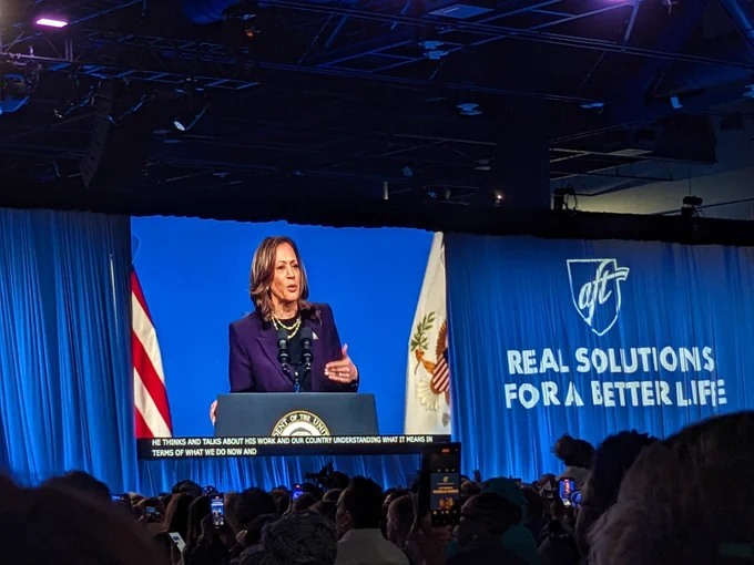 Vice President Kamala Harris appears on a projector screen standing at a podium speaking. On the curtain besides the screen is projected the AFT logo and "Real Solutions for a Better Life". A large crowd is assembled seated before her.