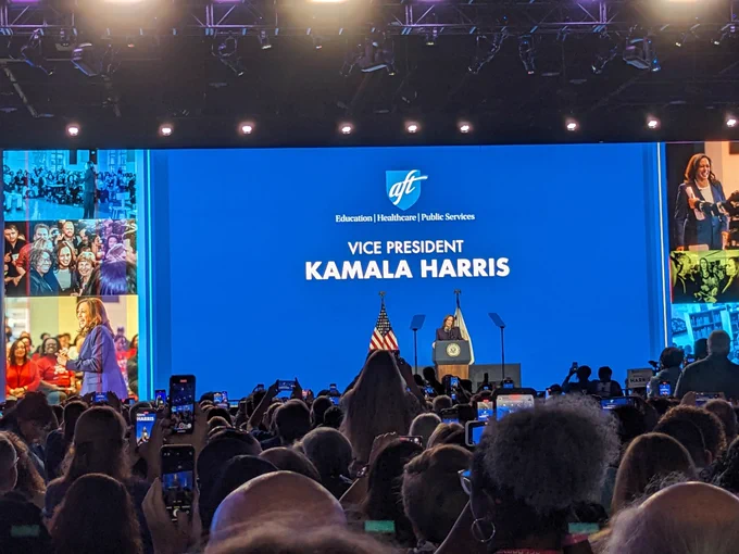 Vice President Kamala Harris stands at a podium on a stage in a large convention hall under the words "Vice President Kamala Harris". She is speaking to a large crowd seated before her.