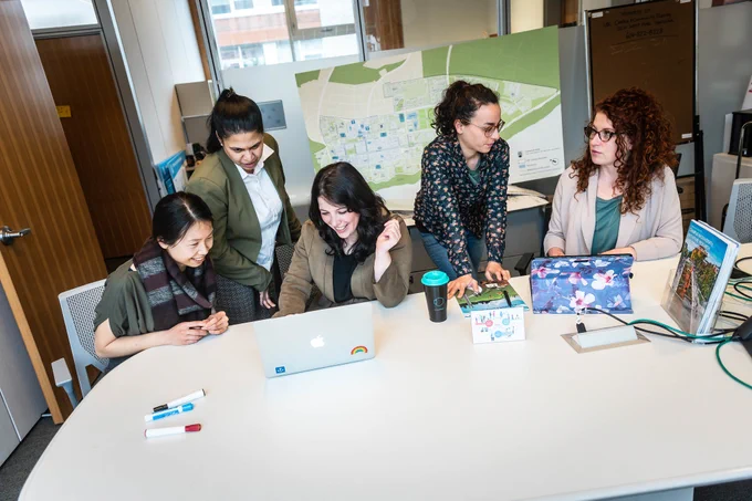 Five staff members working sitting or standing by a table and collaborating together.