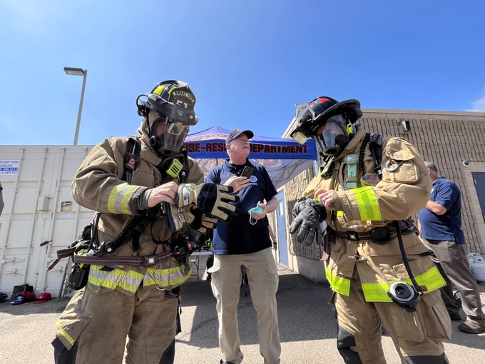 Two firefighters in full gear, including helmets and oxygen tanks, interact outside under a clear sky. 