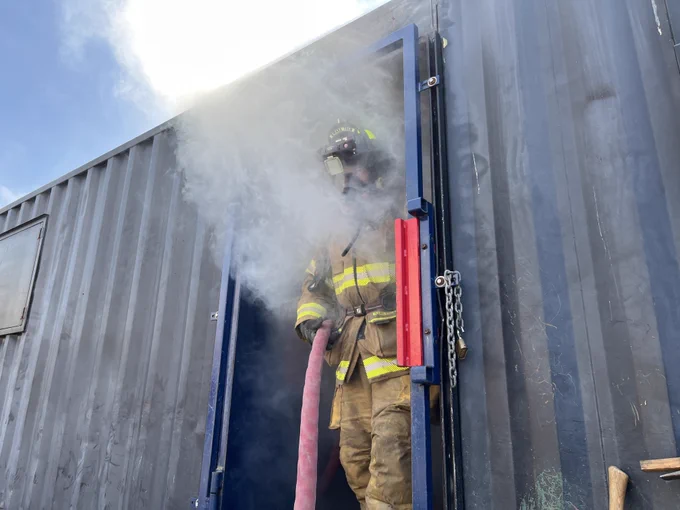 Firefighter in full gear using a hose to spray water on a smoking structure during a daytime operation.