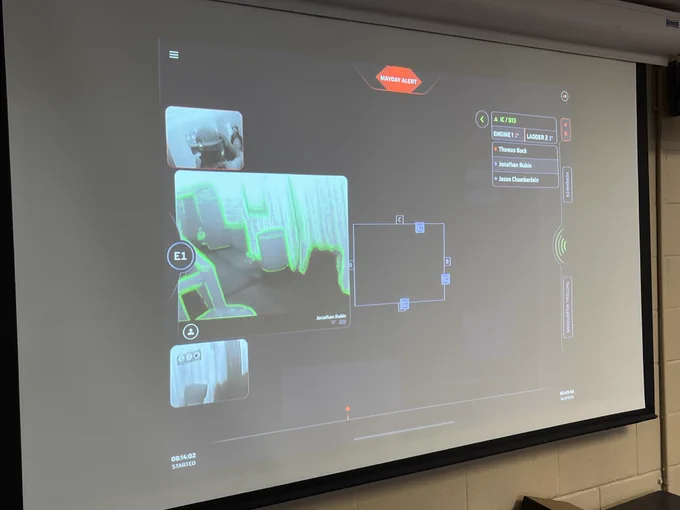 A classroom presentation screen displays an interactive virtual reality interface, with distinct objects labeled, such as a helmet and a chair, enhanced with bright visual aids and annotations in a digital learning environment.