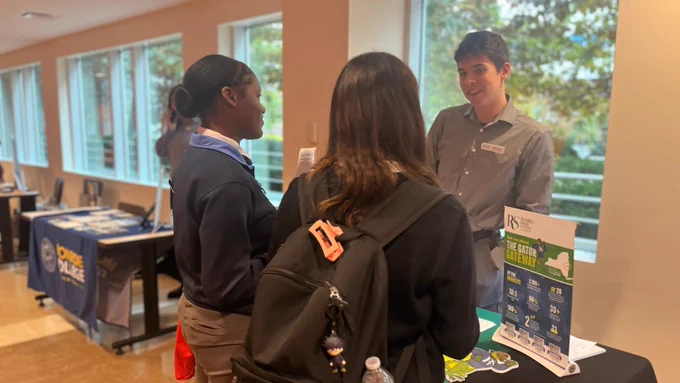 Two students stand and talk to a college representative standing behind a table.