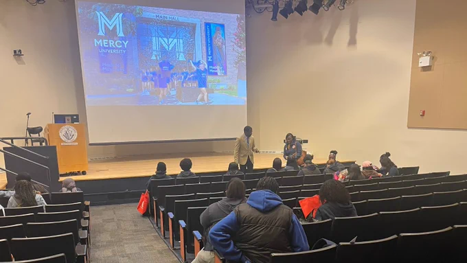Students sit in an auditorium with a screen on a stage that says “Mercy University.”