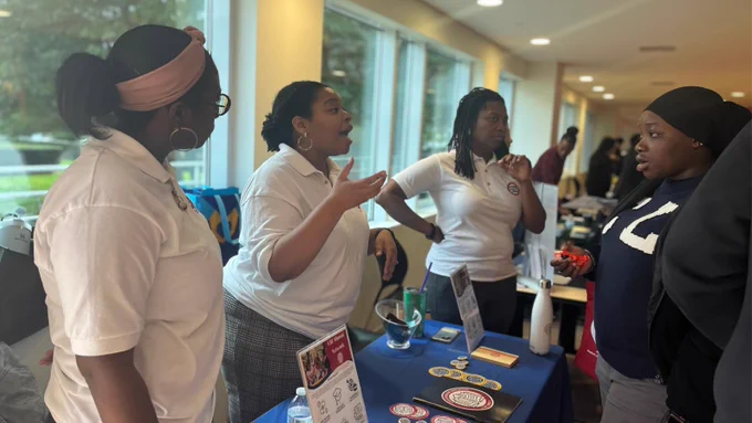 A student stands and talks to three women wearing white t-shirts behind a table. 